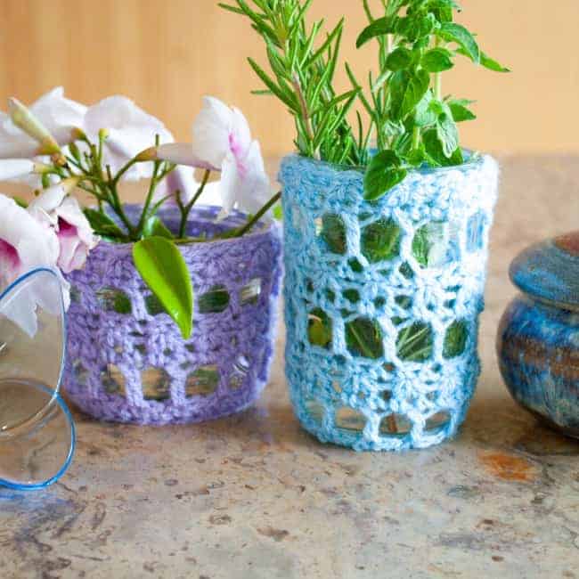 two mason jars with crochet covers in purple and blue holding flowers and greenery
