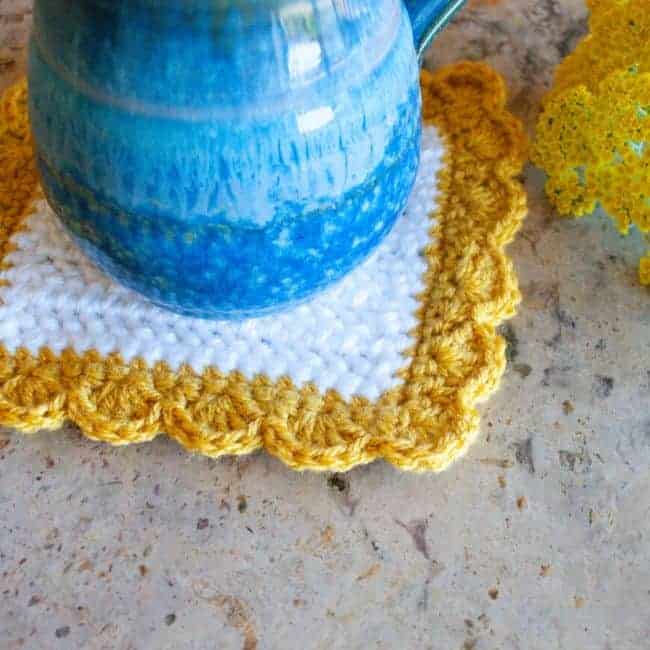 close up of white and yellow square crochet coaster with blue mug