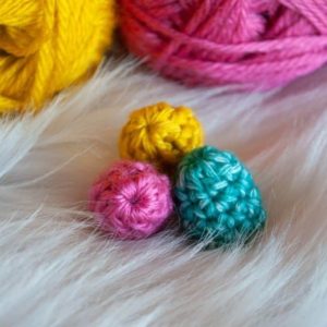 three mini crochet easter eggs in pink, yellow and green in front of two balls of yarn in pink and yellow