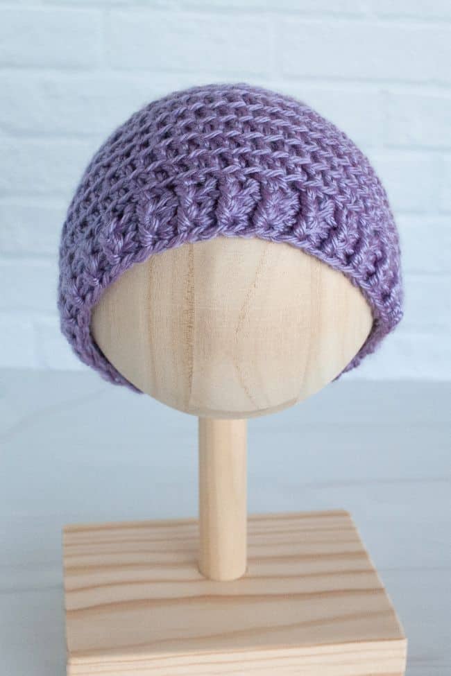 front view of a crochet baby hat in purple on a wooden stand